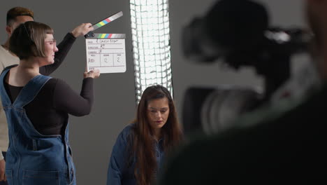 Female-Assistant-On-Film-Set-In-Studio-Using-Clapperboard-Or-Slate-On-Movie-Or-Video-As-Actors-Start-Scene